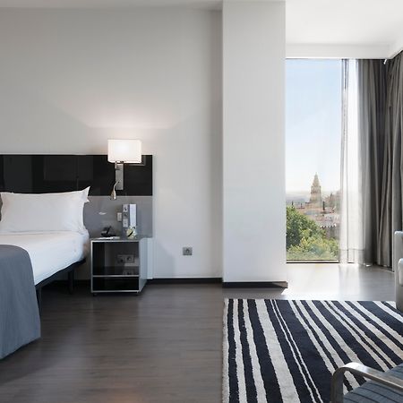 Eurostars Palace Cordoba Luaran gambar The photo shows a modern hotel room featuring a large bed with white linens and dark cushions, paired with a sleek nightstand. There is a contemporary chair and a small table nearby, along with a striped area rug on the floor. A large window allows n