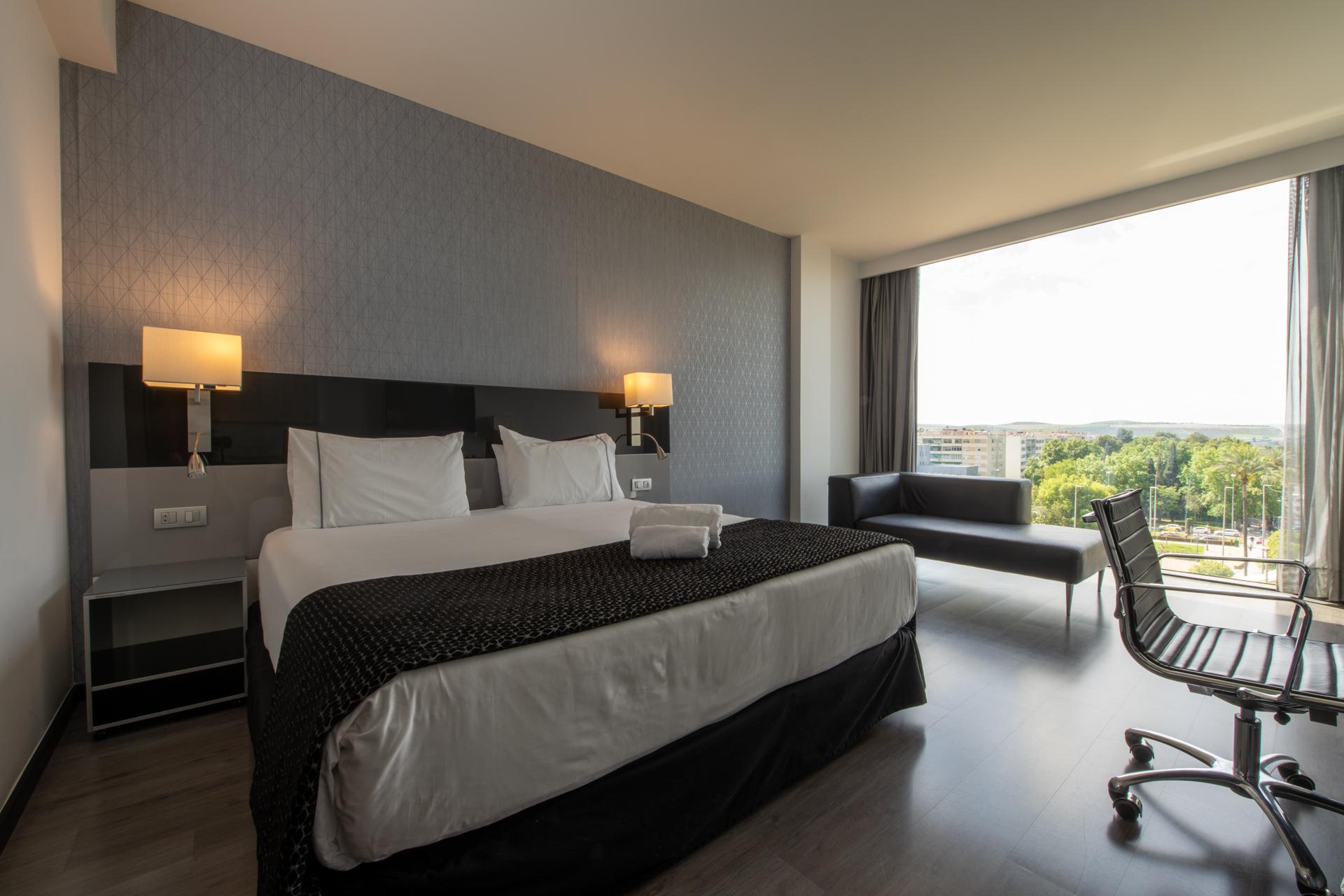 Eurostars Palace Cordoba Luaran gambar The photo shows a modern hotel room featuring a large bed with white linens and a dark patterned throw. On either side of the bed, there are stylish wall-mounted lamps. A contemporary desk chair is positioned at a small desk, and there’s a sofa near 
