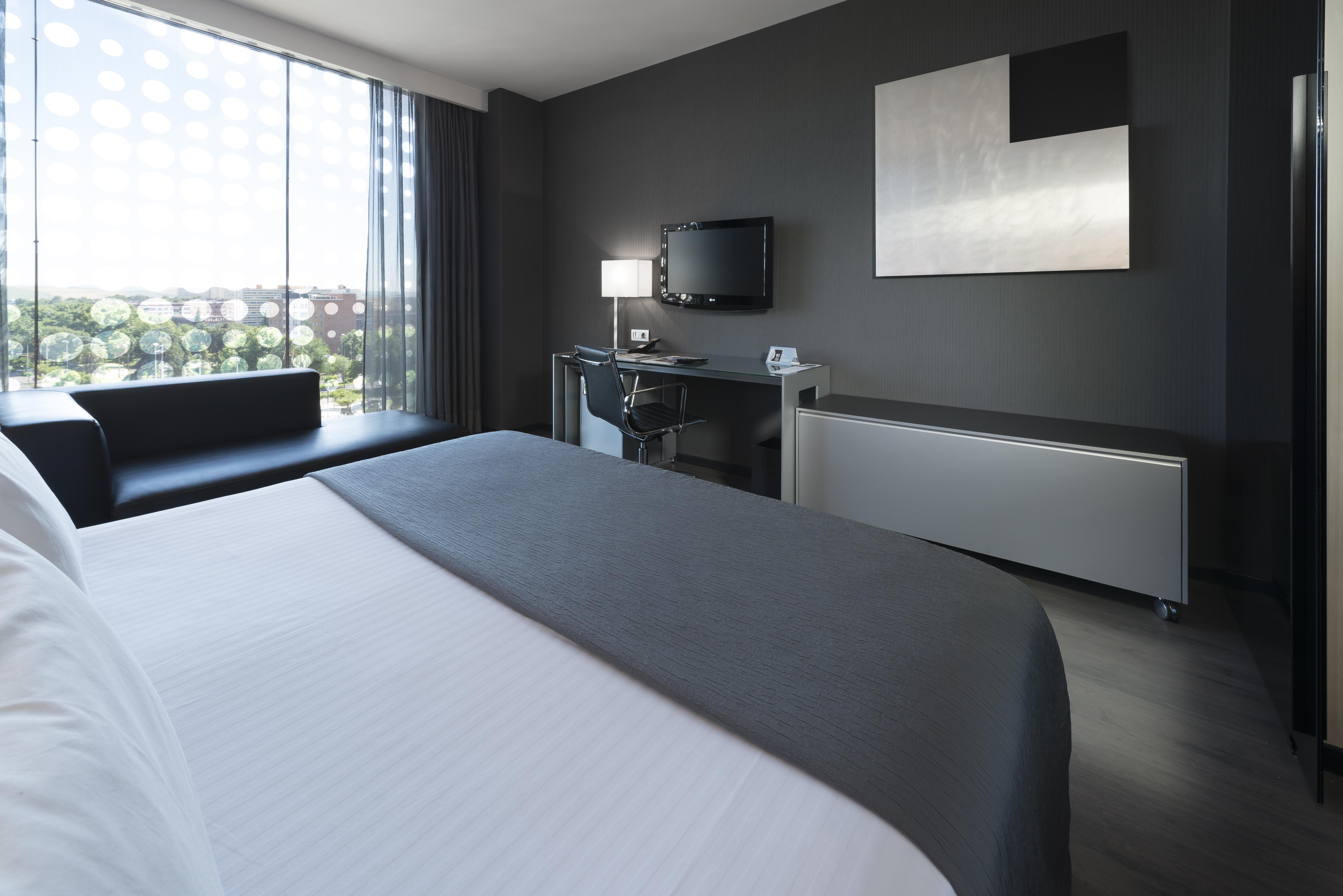 Eurostars Palace Cordoba Luaran gambar The photo shows a modern hotel room featuring a sleek design. In the foreground, there is a bed with a white coverlet and gray accents. To the side, a dark sofa is visible. The room has a large window with sheer curtains, allowing natural light to en