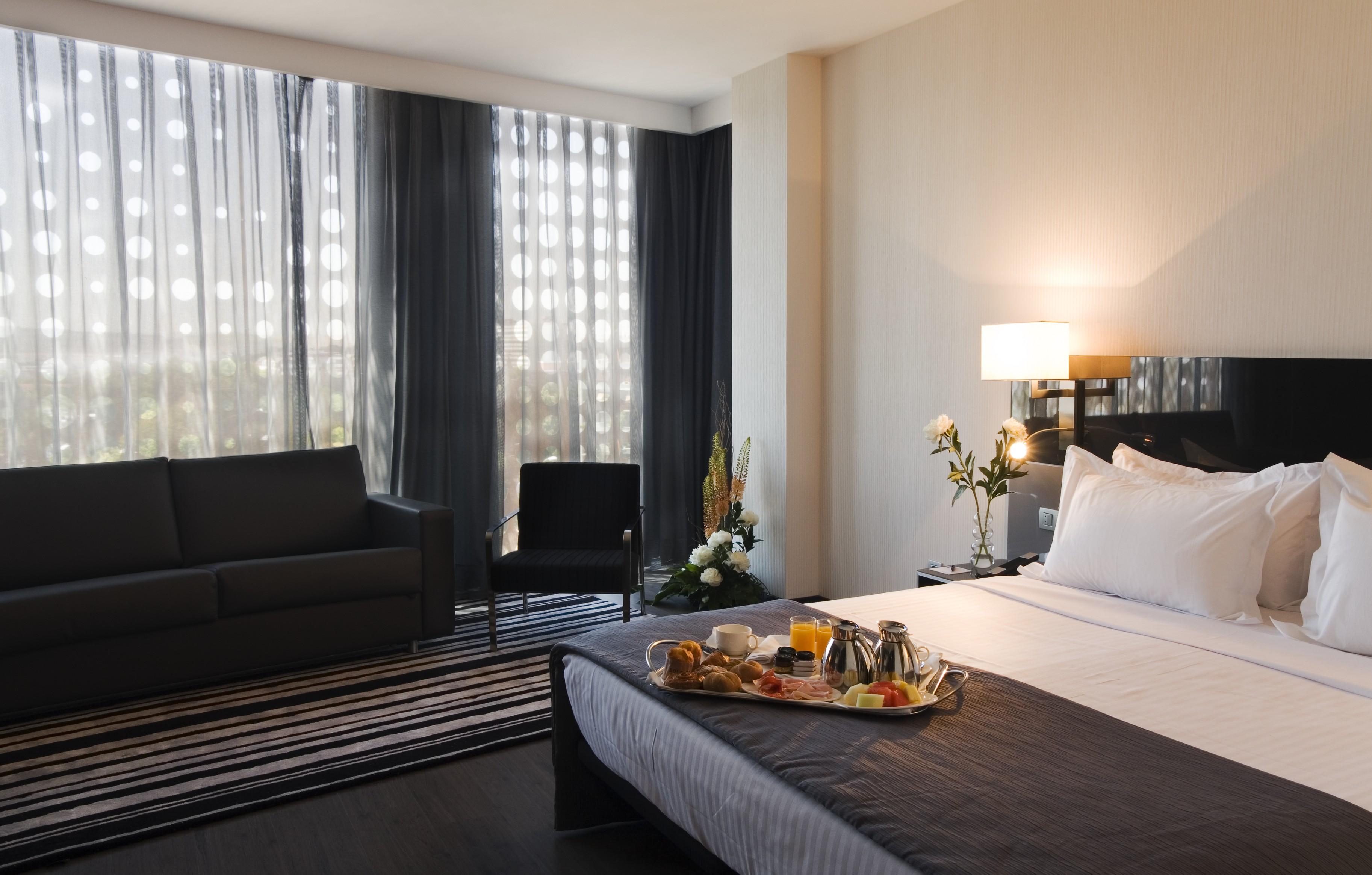 Eurostars Palace Cordoba Luaran gambar The photo shows a modern hotel room featuring a neatly made bed with white linens and a dark grey blanket. There is a tray on the bed with a breakfast spread, including pastries, fruit, and beverages. To the left, there is a comfortable-looking couch
