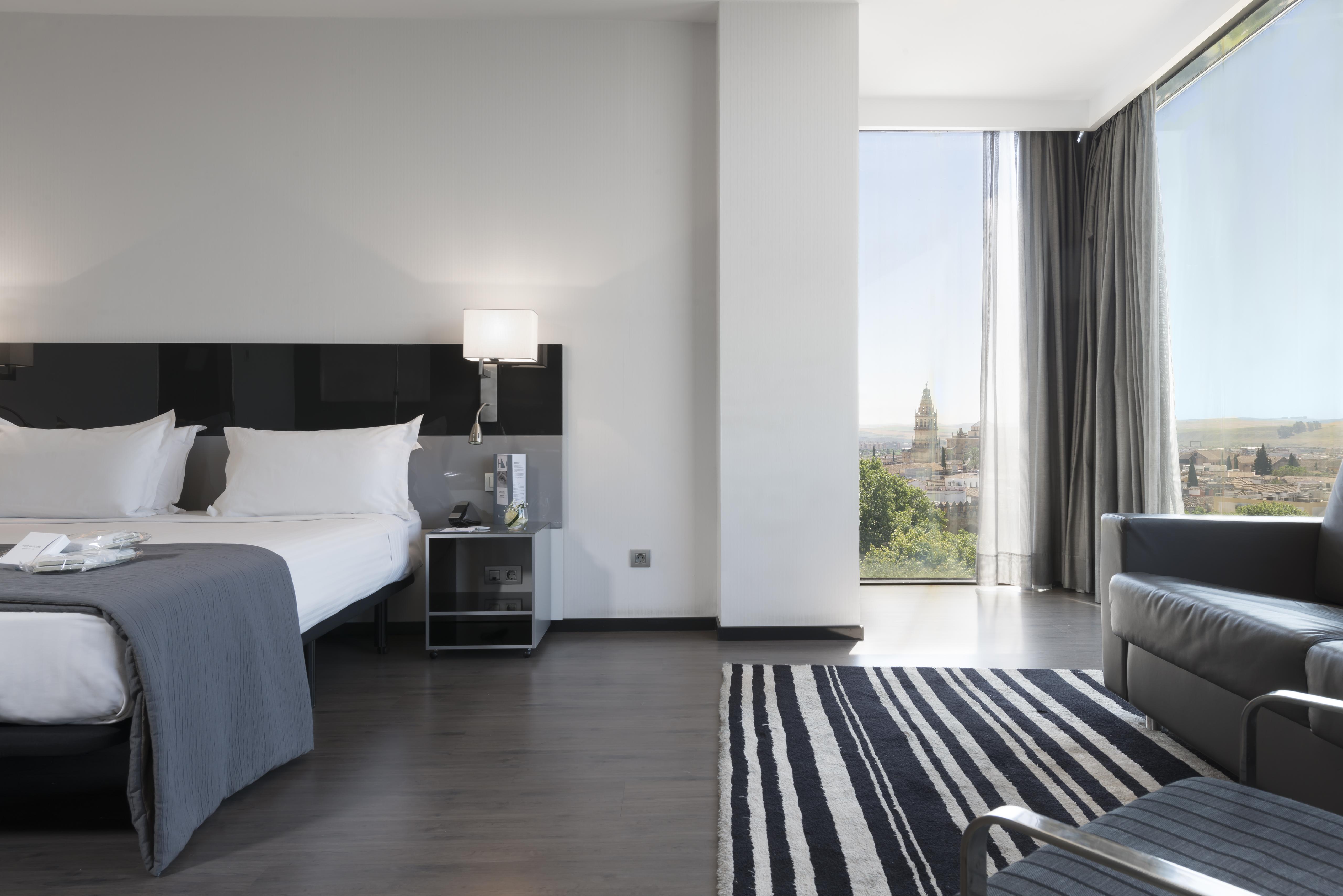 Eurostars Palace Cordoba Luaran gambar The photo shows a modern hotel room featuring a large bed with white linens and dark cushions, paired with a sleek nightstand. There is a contemporary chair and a small table nearby, along with a striped area rug on the floor. A large window allows n
