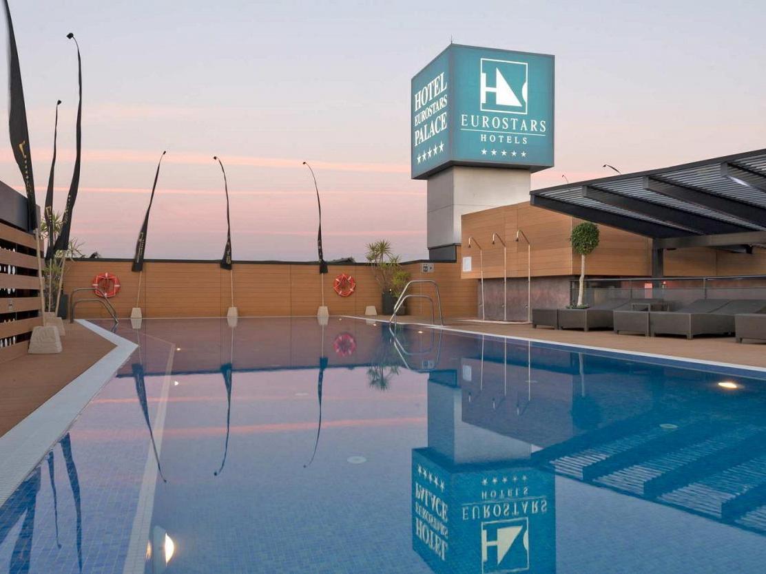 Eurostars Palace Cordoba Luaran gambar The image shows a hotel pool area at twilight. The pool is large and reflective, capturing the colors of the sky and surrounding features. In the background, there is a hotel sign displaying the Eurostars brand, with a modern architecture style. The 