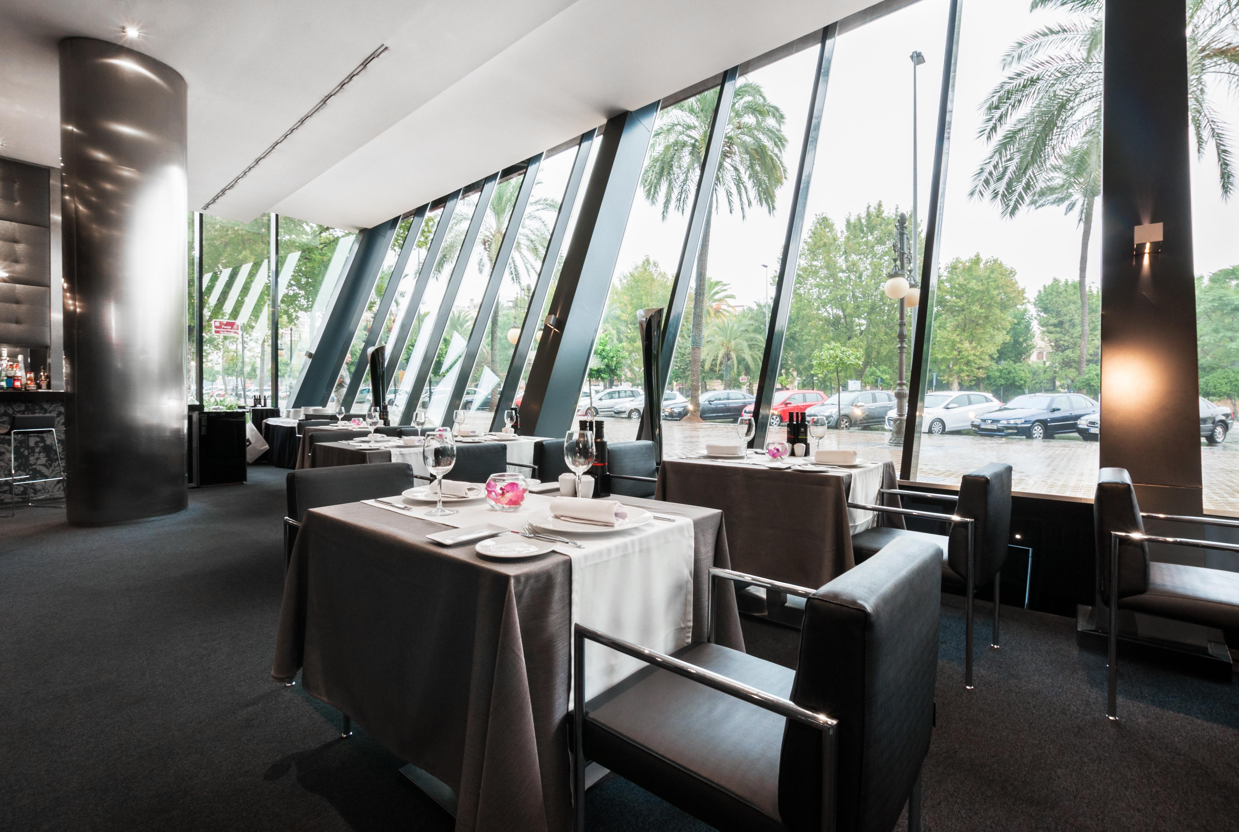 Eurostars Palace Cordoba Luaran gambar The photo shows the interior of a modern restaurant featuring large glass windows that provide an expansive view of the outdoors, including palm trees. The dining area is elegantly set up with tables covered in gray tablecloths, decorated with simple