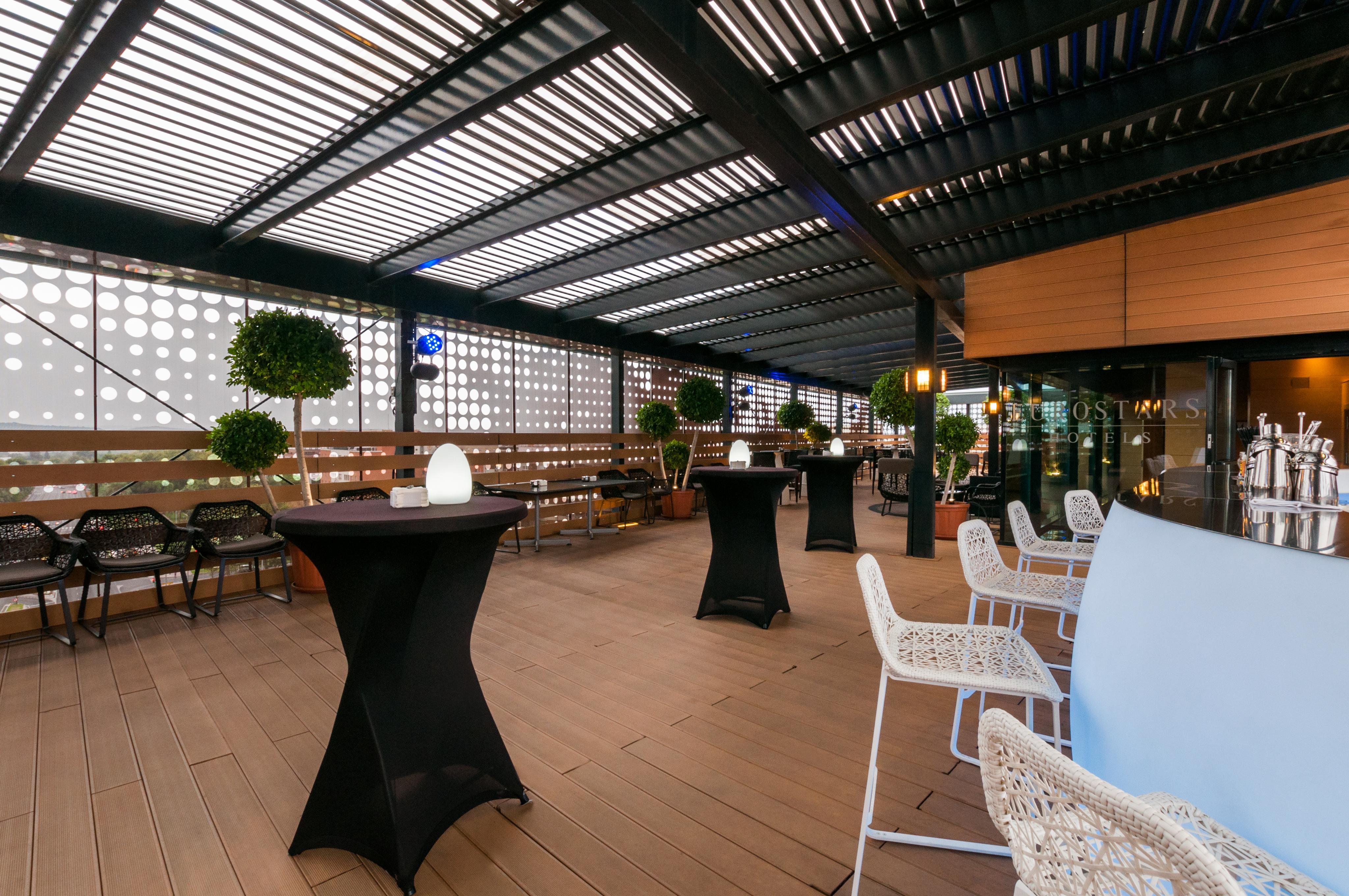 Eurostars Palace Cordoba Luaran gambar The photo shows a stylish outdoor seating area, likely part of a restaurant or lounge. It features a wooden floor and an overhead structure with horizontal slats, allowing for both shade and light. There are several tall, black cocktail tables and wh