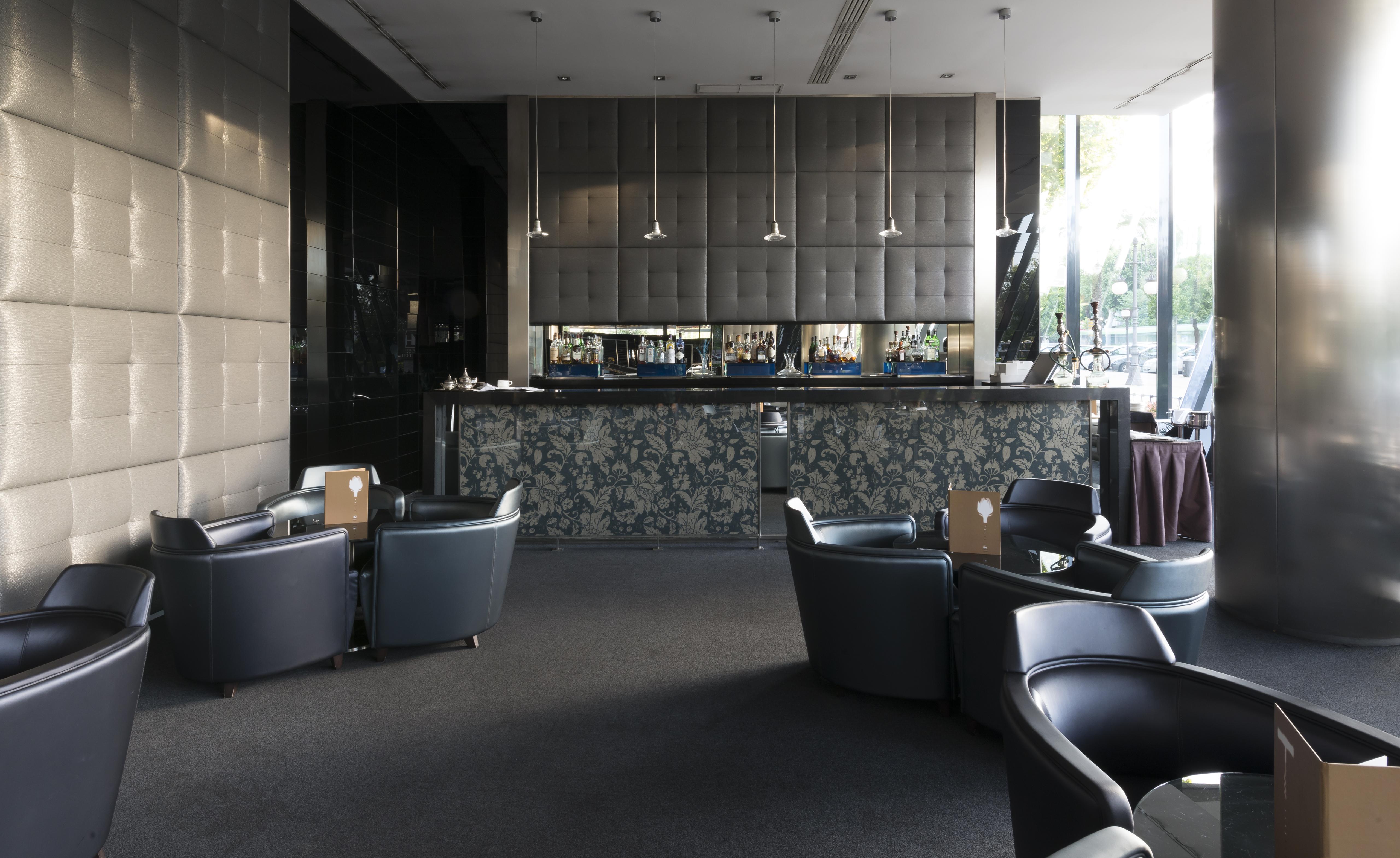 Eurostars Palace Cordoba Luaran gambar The photo shows a modern bar or lounge area. In the foreground, there are several comfortable black armchairs arranged around small tables. The background features a sleek bar counter with a dark, textured wall behind it. The bar is illuminated with 