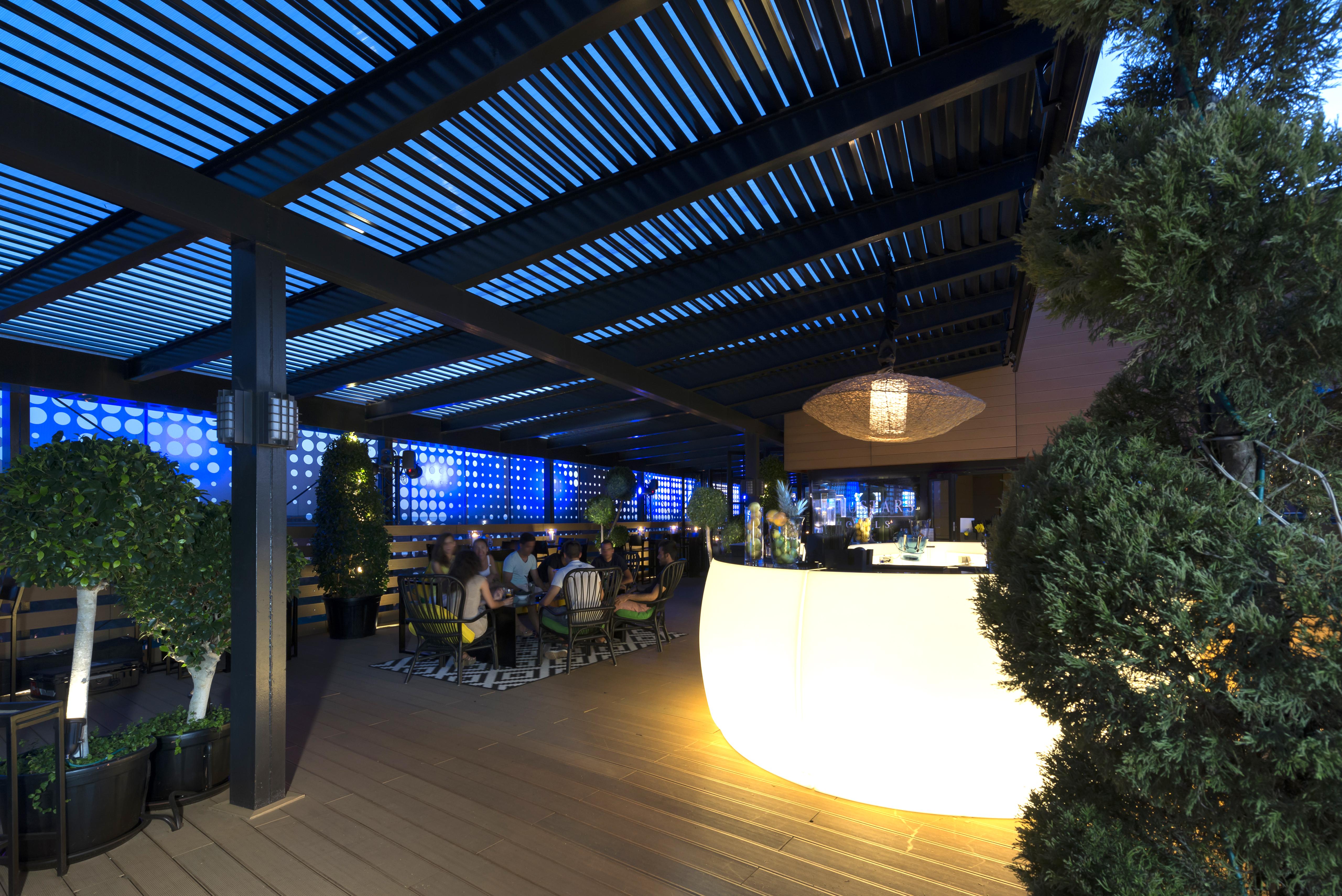 Eurostars Palace Cordoba Luaran gambar The image shows a modern outdoor bar or restaurant area. The space features a sleek, illuminated bar that stands out with its curved shape. Patrons can be seen seated around tables, enjoying the ambiance. The overhead structure consists of slatted pa