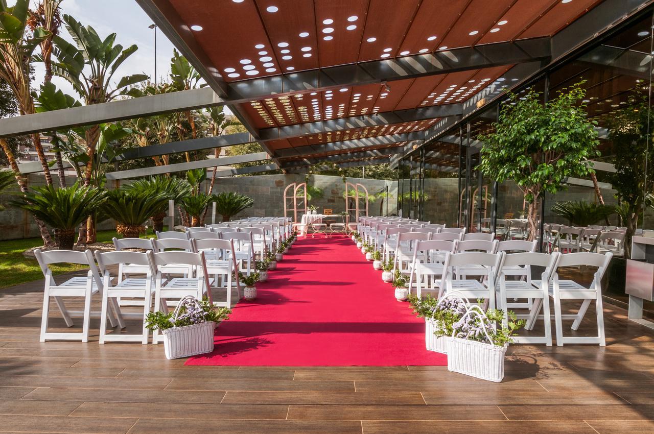 Eurostars Palace Cordoba Luaran gambar The image shows an outdoor wedding setup. There are white chairs arranged neatly on either side of a vibrant red carpet that leads to a framed archway. The area is covered by a glass and metal structure, allowing plenty of natural light, while tropic