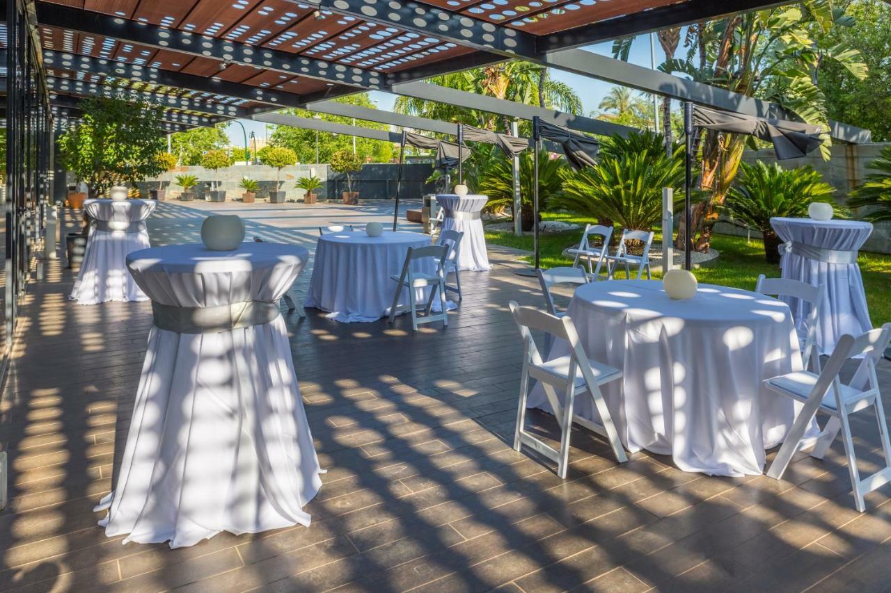 Eurostars Palace Cordoba Luaran gambar The photo shows an outdoor event setup. There are several round tables covered with white tablecloths, some adorned with decorative elements like small centerpieces. Chairs are arranged around the tables, and the area is surrounded by greenery, inclu